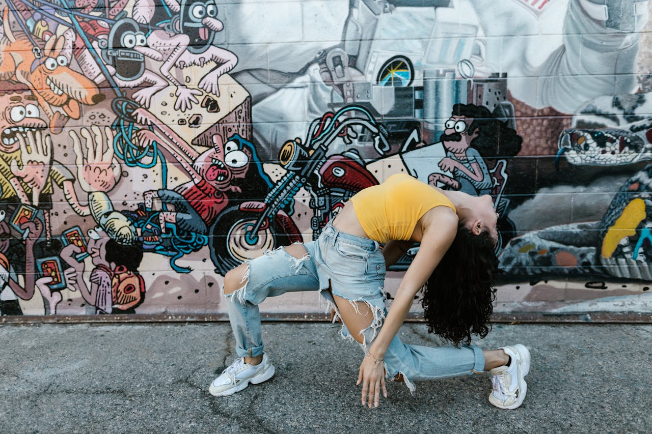 A woman gracefully bends in denim jeans against a vibrant graffiti wall.